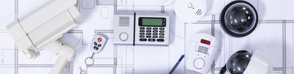 Security devices including CCTV cameras, a keypad, a smoke detector, a remote control, and a motion sensor are arranged on top of architectural blueprints, highlighting the importance of security and safety in building design. by Standex Detect