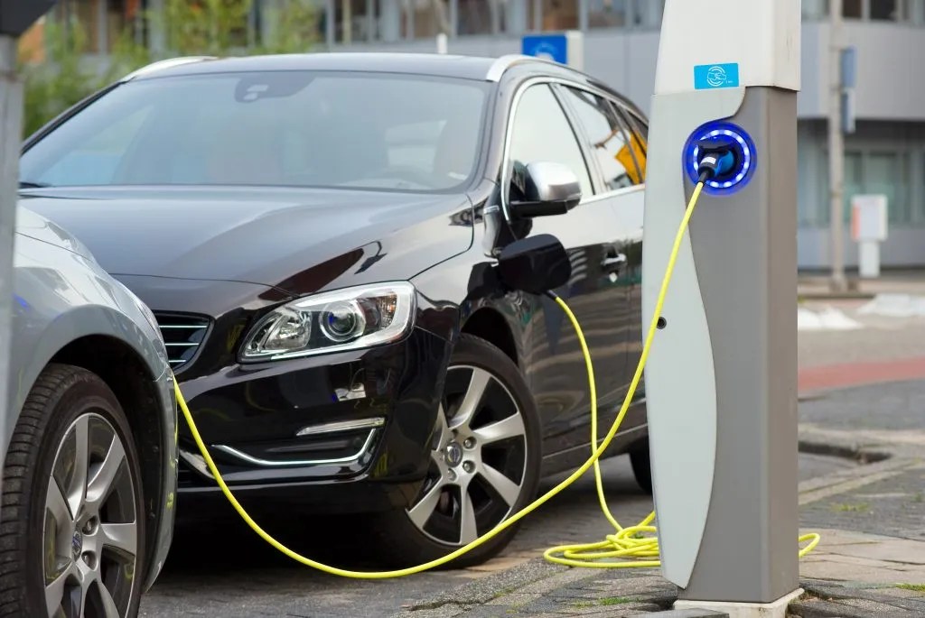 A black E-Car is parked on the street and plugged into a charging station with a yellow cable. The blue-lit station ensures safe charging through E-Car Isolation, while another car is parked nearby. by Standex Detect