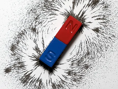 A red and blue bar magnet labeled N and S lies on paper, surrounded by black iron filings that reveal the magnetic field pattern and demonstrate the basic principles of magnets. by Standex Detect
