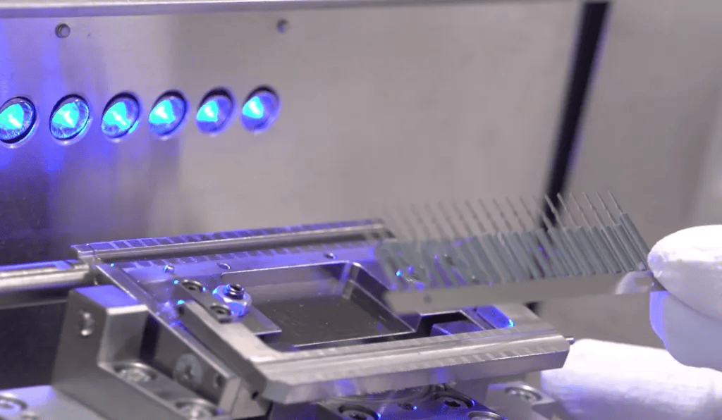 A gloved hand holds a glass slide with etched lines near a metallic device, illuminated by blue LED lights in a laboratory setting, illustrating how a reed switch works in precision instrumentation. by Standex Detect