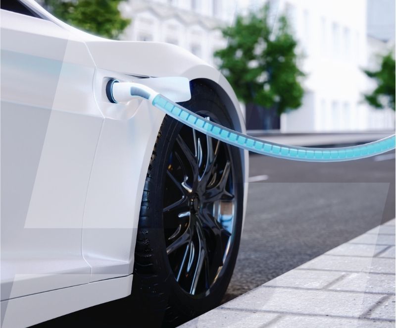 Close-up of a white EV charging at a street-side station, with the charging cable plugged in near the front wheel. Reed relays and high-voltage measurement enable safe and efficient operation. Trees and buildings are blurred in the background. by Standex Detect