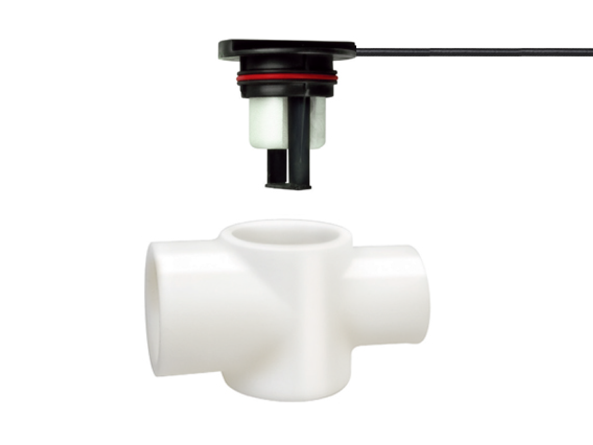 A black and white FPS-T Series electronic sensor with a cable is shown above a white plastic pipe fitting with four openings arranged in a cross shape, both against a plain white background, ideal for HVAC or liquid level sensor applications. by Standex Detect